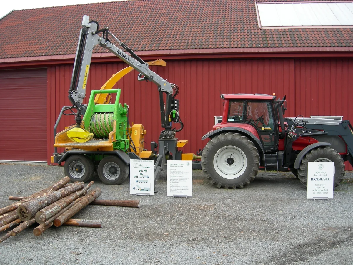 Traktor med flishogger og stablet tømmer foran den røde låven, med skilt for biodiesel.